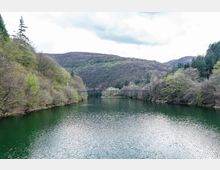 Широко езеро или язовир, заобиколено от гористи хълмове, с зелена вода и леко вълнение на повърхността. В далечината се вижда тесен мост, пресичащ водата, под облачно небе.