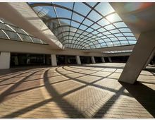 Покрит вътрешен двор в модерна сграда с голямо извито стъклено покритие на метална решетка, което пропуска слънчева светлина и хвърля геометрични сенки върху настилка от тухлени павета. Пространството е обградено от наклонени колони и редица остъклени входове по периметъра.