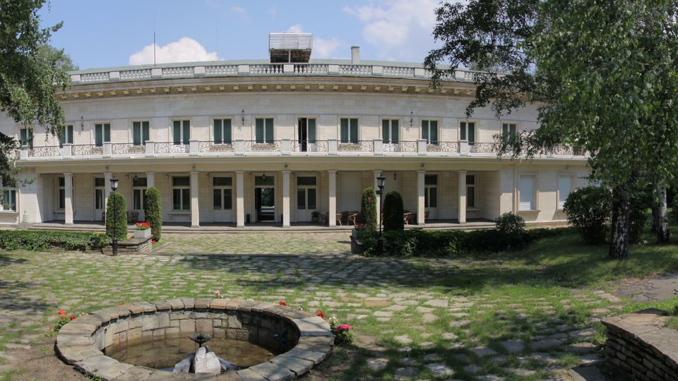 Широка, двуетажна светла сграда с дълга фасада, балкон с декоративни парапети и покрита колонада на приземния етаж, разположена в парк. Пред нея има каменна площадка и кръгъл каменен фонтан, заобиколени от тревни площи и високи дървета.