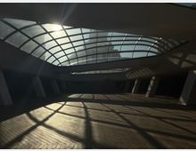 Вътрешно пространство на съвременна сграда с голям извит стъклен покрив на метална решетка, през който навлиза слънчева светлина. Подът е с павирани плочки, а наклонени колони и сенки от покрива оформят геометрични шарки по настилката.