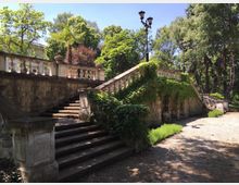 Каменни стълби в парк водят към тераса с балюстрада и декоративни вази, като част от парапета е покрита с увивни растения. Наоколо има високи дървета и алея, осветена с класически улични лампи.