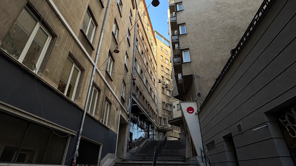 Тясна градска улица между високи жилищни сгради с множество прозорци, заснета отдолу нагоре. В центъра има широка стълба с парапети, водеща към по-високо ниво, с окачени улични лампи и видимо небе над сградите.