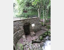 A stone structure with an arched entrance sits next to a small stream flowing over moss-covered rocks in a lush forest setting. The area is surrounded by trees and features a metal railing along the top of the structure for safety.