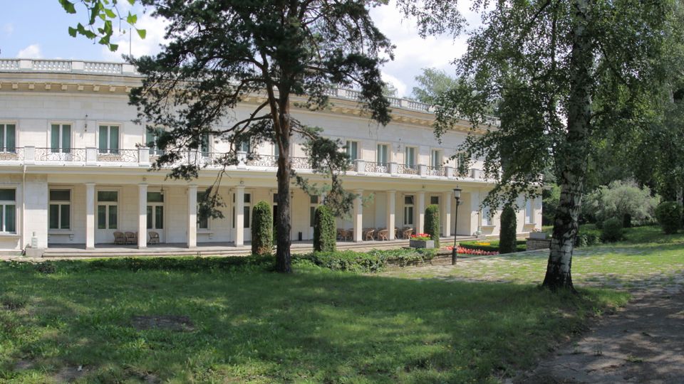 Дълга двуетажна бяла сграда с класическа фасада, колонада и балкони с декоративни метални парапети. Отпред има тревна площ с високи дървета, алея и малки цветни лехи в паркова среда.