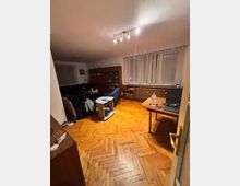 Interior living room with herringbone parquet wood flooring, a blue sofa and armchair around a small coffee table, and a tall bookcase along the back wall. Large windows with sheer curtains line the right side above a radiator cover, with a simple ceiling track light and a small desk near the doorway.