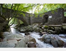 A stone bridge with an arched design spans over a flowing rocky stream, surrounded by lush green trees. The bridge features a small doorway and steps on the right side, with moss-covered rocks and natural vegetation enhancing the setting.