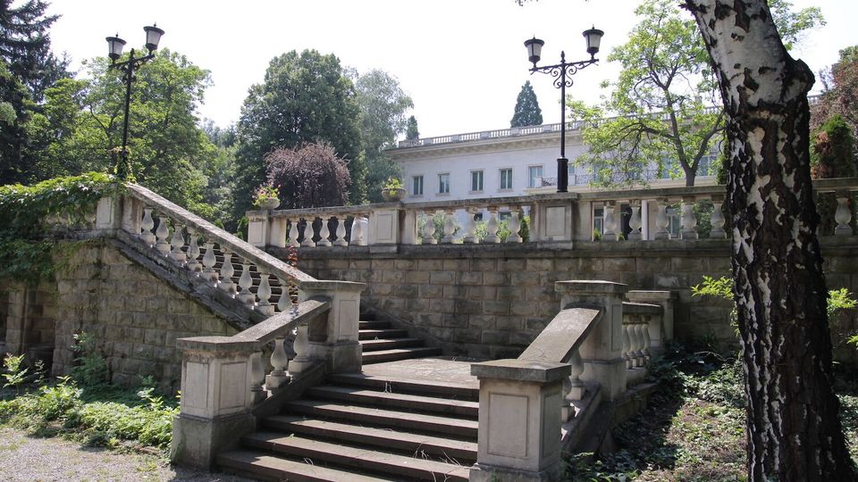 Каменни стълби в парк, с парапети от балюстри и тераса със зидана ограда отгоре. Около тях има високи дървета и декоративни улични лампи, а на заден план се вижда светла класическа сграда.