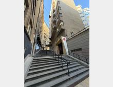 Стръмно външно стълбище с метални парапети в тясна градска уличка между високи жилищни блокове с балкони и прозорци. В подножието има вход към сграда и стена с наклонена рампа, а над тях се вижда ясно синьо небе.