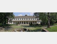 Широка, двуетажна светла сграда с дълга фасада, балкон с декоративни парапети и покрита колонада на приземния етаж, разположена в парк. Пред нея има каменна площадка и кръгъл каменен фонтан, заобиколени от тревни площи и високи дървета.