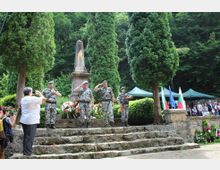 A stone memorial surrounded by tall evergreen trees and lush greenery, featuring a vertical obelisk-style monument adorned with a decorative emblem. The site includes stone steps leading to the base of the monument, flanked by flags and tents in the background.