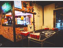 Интериор на бар с тухлени стени, неонови табели и винтидж плакати, с отвор към барплота и осветени хладилници. На преден план има маса за бирпонг с подредени червени пластмасови чаши, а вдясно се вижда входна врата с остъкление.