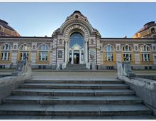 Фасада на голяма историческа обществена сграда с симетрична композиция, жълто-бели хоризонтални ивици и аркадни прозорци. В центъра има монументален вход с висока арка и голямо остъклено прозоречно поле над стълбище, а отстрани се виждат куполни покриви и декоративни орнаменти.