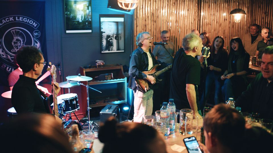 Интериор на малък бар с нисък таван, сцена за жива музика с барабани, китари и микрофон, осветена с цветни сценични светлини. По стените има дървена ламперия и плакати, а отпред се виждат маси с чаши и бутилки и тясно пространство за публика.