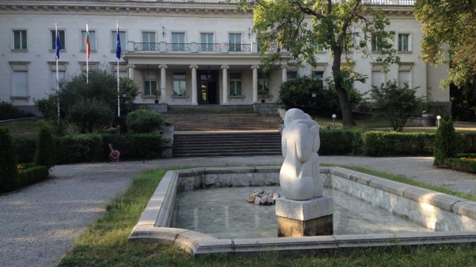 Бяла неокласическа обществена сграда с централен портик и колони, пред която има стълбище и три флагщока. На преден план се вижда правоъгълен каменен басейн с плитка вода и скулптура в средата, заобиколен от алеи и зеленина.