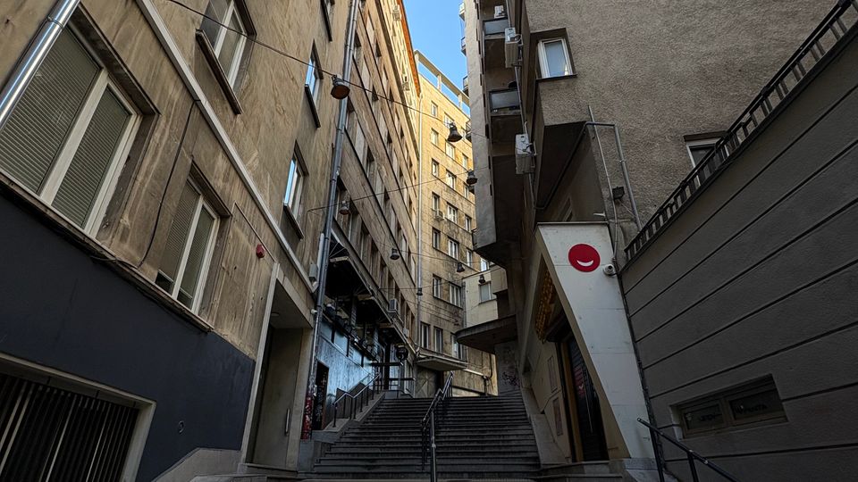 Стръмно градско стълбище с метален парапет в средата, разположено в тясна уличка между високи жилищни сгради с прозорци и балкони. Погледът е насочен нагоре към светлото небе, а по фасадите се виждат улични лампи и тръби.