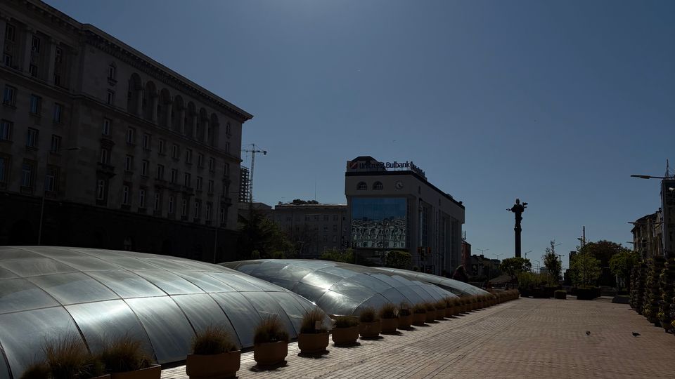 Широка градска пешеходна алея с настилка от павета и редица саксии, до която се простират ниски полукръгли стъклени куполи като покрив на подземно пространство. В далечината се виждат високи административни сгради и монументална колона на площад, под силно слънце в ясно небе.