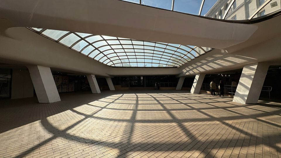 Широко закрито градско пространство с тухлена настилка и голям извит стъклен покрив, който пропуска дневна светлина и хвърля решетъчни сенки на пода. Зоната е оформена с масивни наклонени колони, парапети по периферията и фасади на сгради, видими през остъкляването.