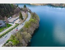 Въздушен изглед към извита двулентова пътна отсечка покрай брега на голямо синьо езеро. От едната страна има гориста ивица и малък паркинг до сграда, а от другата – спокойна водна повърхност по крайбрежието.