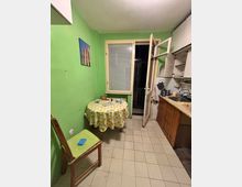 Small kitchen with green painted walls, a tiled floor, and a round table with a patterned tablecloth beside a window with blinds. To the right is a countertop with sink and white upper cabinets, and an open door leading to a dark balcony or exterior space.