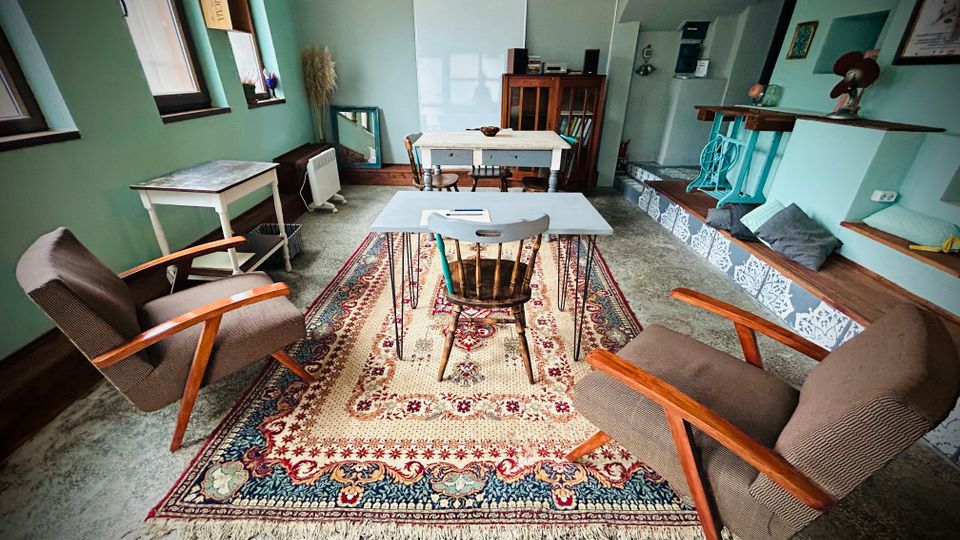 Интериор на малка всекидневна или общо помещение с зелени стени, дълъг прозоречен ред и килим в центъра с ниска маса и два ретро фотьойла. В дъното има бяла дъска и дървен шкаф, а вдясно — повдигната платформа със стъпала с шарени плочки, бар плот и вградени места за сядане с възглавници.