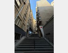 Стръмно градско стълбище с метален парапет в средата, разположено в тясна уличка между високи жилищни сгради с прозорци и балкони. Погледът е насочен нагоре към светлото небе, а по фасадите се виждат улични лампи и тръби.