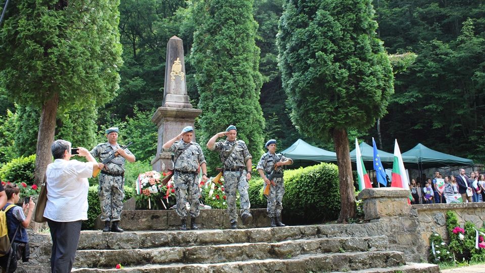A stone memorial surrounded by tall evergreen trees and lush greenery, featuring a vertical obelisk-style monument adorned with a decorative emblem. The site includes stone steps leading to the base of the monument, flanked by flags and tents in the background.