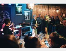 Интериор на малък бар с нисък таван, сцена за жива музика с барабани, китари и микрофон, осветена с цветни сценични светлини. По стените има дървена ламперия и плакати, а отпред се виждат маси с чаши и бутилки и тясно пространство за публика.