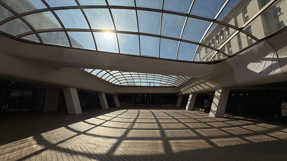 Покрит вътрешен двор или пасаж с голям извит стъклен покрив на метална решетка, през който влиза силна дневна светлина. На пода от павета се виждат решетъчни сенки, а пространството е обградено от масивни колони и фасади на сгради отстрани.