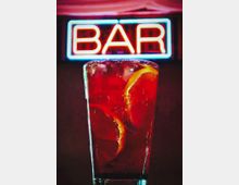 Неонова табела с надпис „BAR“ на тъмен фон, заснета в близък план. На преден план има висока чаша с лед и напитка, гарнирана с резен портокал.