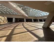 Покрито атриумно пространство с извита стъклена покривна конструкция и масивни наклонени колони, над настилка от тухлени павета. Вляво има открити каменни руини, а в дъното се вижда редица стъклени входове и фасада на сгради през покрива.