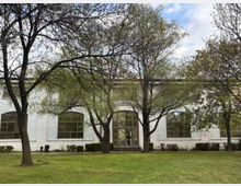 Едноетажна светла сграда с големи арковидни прозорци и централна остъклена врата със стълби, разположена зад тревна площ. Пред фасадата има няколко високи дървета с разперени клони и ниски храсти покрай стената.