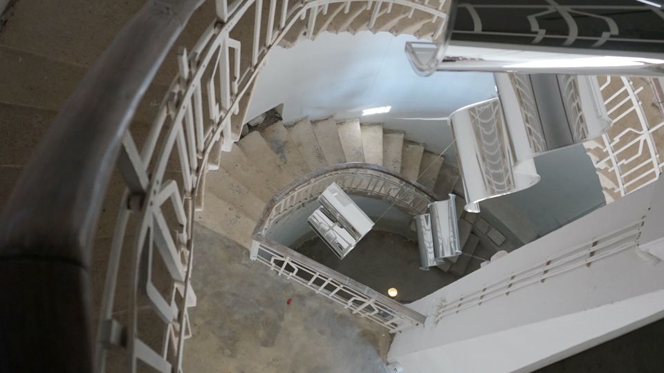 Гледка отгоре към тясно спираловидно стълбище с бетонни стъпала и бели метални парапети, спускащо се през няколко нива. Стълбищната клетка е с бели стени и малка площадка на дъното.