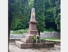 A stone monument shaped like an obelisk with inscriptions and a golden emblem, surrounded by evergreen trees and set against a low stone wall and dense greenery. A floral wreath is placed at its base, and the ground is paved with stone.