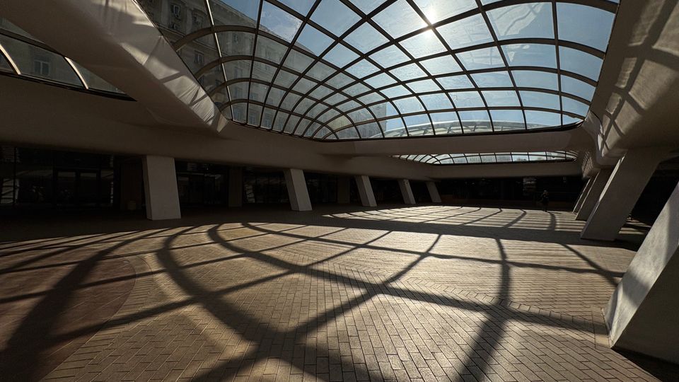 Широко вътрешно атриумно пространство в модерна сграда с извита стъклена покривна конструкция на метална решетка. Слънчевата светлина през покрива хвърля мрежести сенки върху тухлената настилка и белите наклонени колони около периметъра.