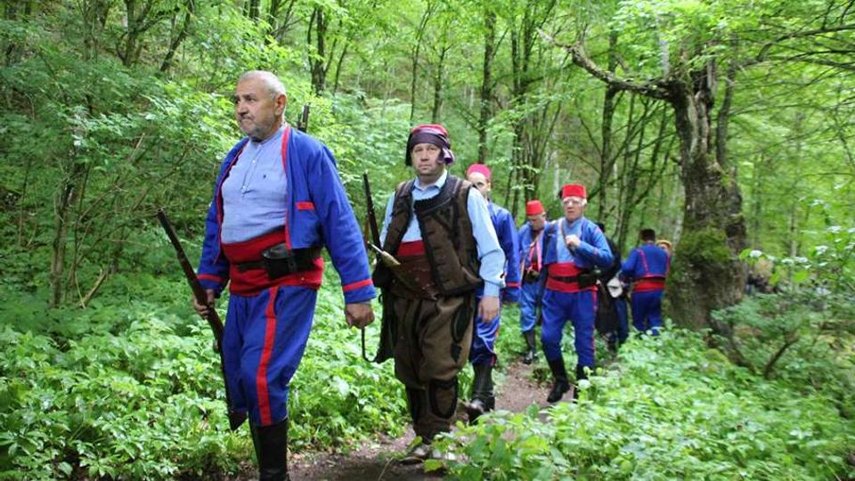 A group of individuals walking along a narrow dirt path in a dense forest with vibrant green foliage and mature trees. The setting features abundant undergrowth and a natural, shaded environment.