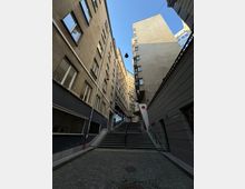 Тясна градска улица между високи жилищни сгради с множество прозорци, заснета отдолу нагоре. В центъра има широка стълба с парапети, водеща към по-високо ниво, с окачени улични лампи и видимо небе над сградите.