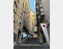 Тясна градска уличка между високи жилищни сгради, с широка стълба и метални парапети, водещи нагоре. Над стълбите има окачени улични лампи на кабели, а фасадите са с прозорци, балкони и външни климатични тела.