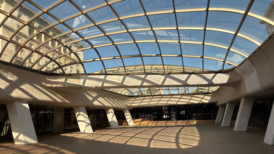 Покрито градско пространство с голям извит стъклен покрив в метална решетка, през който се вижда синьо небе и околни сгради. Под него има широка павирана площ с колони, входове към сграда и силни светлосенки от конструкцията върху пода.