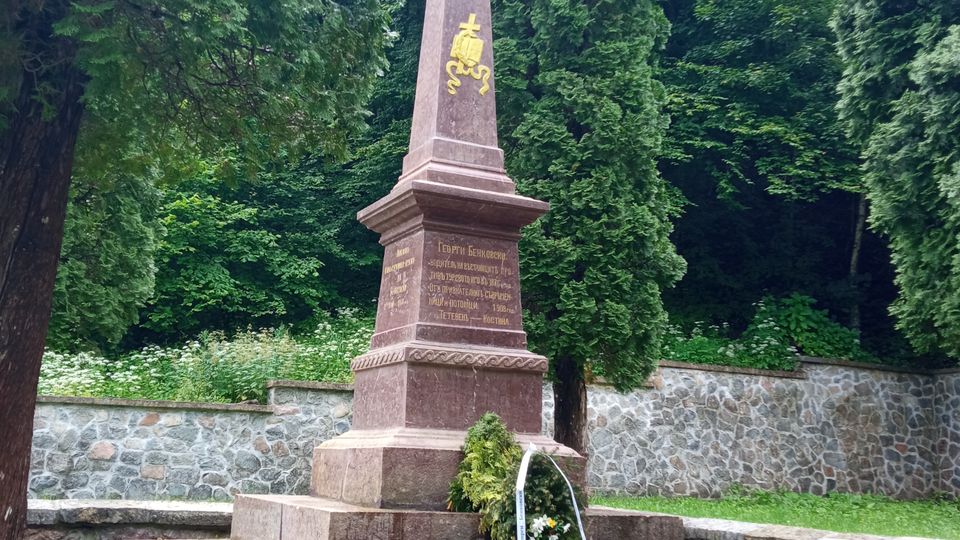 A stone monument shaped like an obelisk with inscriptions and a golden emblem, surrounded by evergreen trees and set against a low stone wall and dense greenery. A floral wreath is placed at its base, and the ground is paved with stone.
