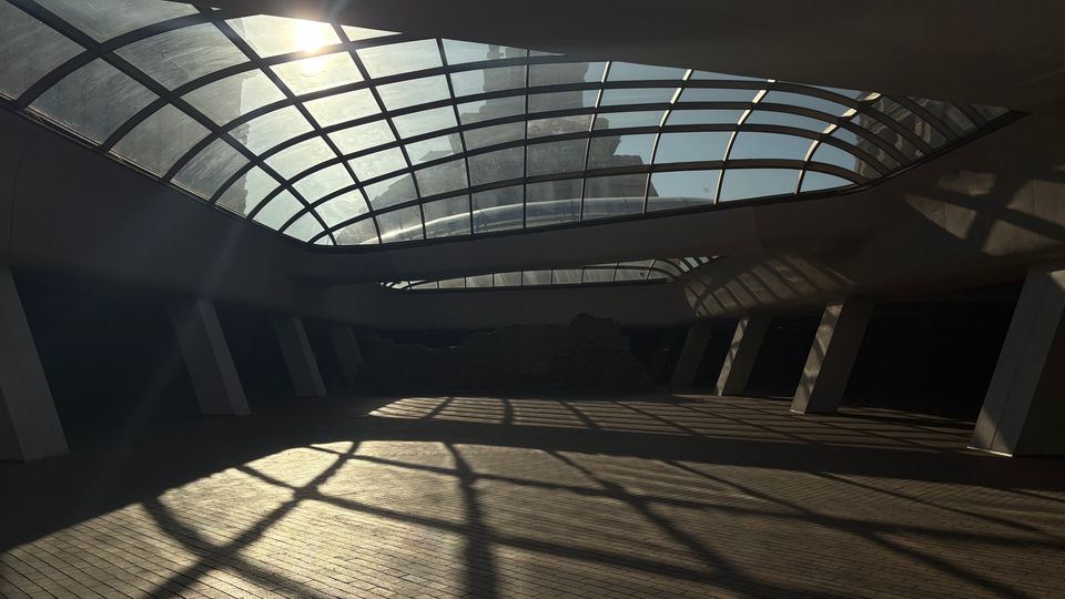 Вътрешно пространство на съвременна сграда с голям извит стъклен покрив на метална решетка, през който навлиза слънчева светлина. Подът е с павирани плочки, а наклонени колони и сенки от покрива оформят геометрични шарки по настилката.