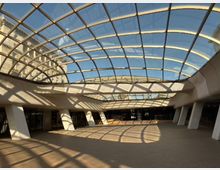 Покрито градско пространство с голям извит стъклен покрив в метална решетка, през който се вижда синьо небе и околни сгради. Под него има широка павирана площ с колони, входове към сграда и силни светлосенки от конструкцията върху пода.