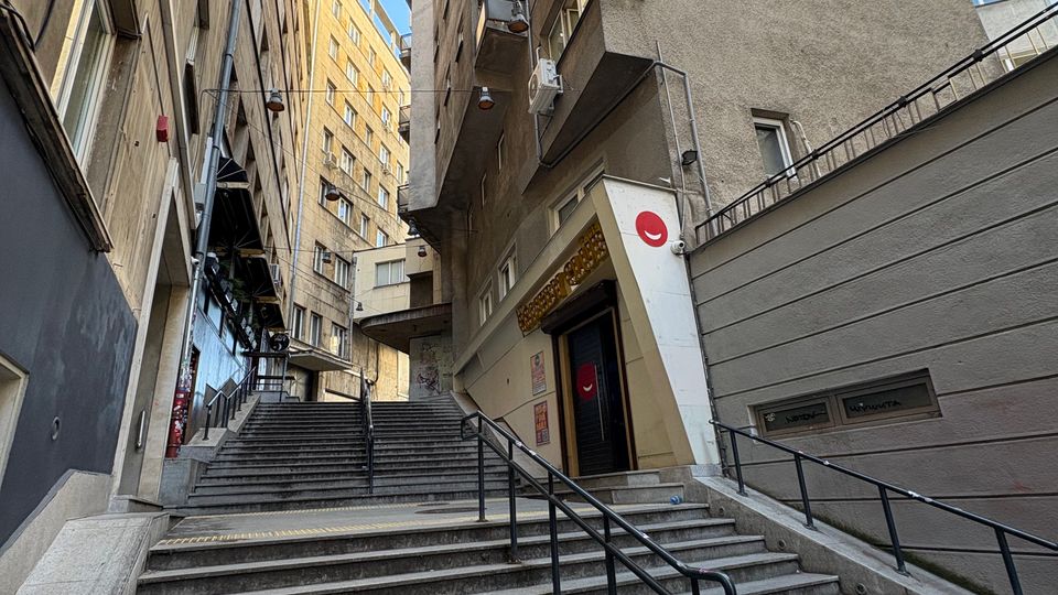 Стръмно външно стълбище с метални парапети в тясна градска уличка между високи жилищни блокове с балкони и прозорци. В подножието има вход към сграда и стена с наклонена рампа, а над тях се вижда ясно синьо небе.