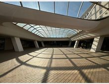 Широко закрито градско пространство с тухлена настилка и голям извит стъклен покрив, който пропуска дневна светлина и хвърля решетъчни сенки на пода. Зоната е оформена с масивни наклонени колони, парапети по периферията и фасади на сгради, видими през остъкляването.