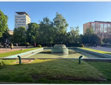 Градски парк с правоъгълен плитък басейн и декоративен фонтан в центъра, ограден от тревни площи и алеи. На заден план се виждат високи сгради, дървета и открито небе.