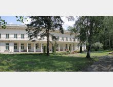 Дълга двуетажна бяла сграда с класическа фасада, колонада и балкони с декоративни метални парапети. Отпред има тревна площ с високи дървета, алея и малки цветни лехи в паркова среда.