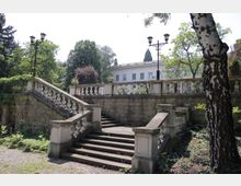 Каменни стълби в парк, с парапети от балюстри и тераса със зидана ограда отгоре. Около тях има високи дървета и декоративни улични лампи, а на заден план се вижда светла класическа сграда.