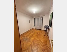 Empty interior room with white walls and a herringbone-pattern hardwood floor, lit by a ceiling light. A window with sheer curtains and blinds is centered on the far wall, with minimal wall art and a small stand near the right wall.