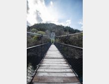 Висящ пешеходен мост с дървена настилка и метални мрежести парапети, водещ към бяла метална конструкция в средата. Мостът преминава над гориста долина с хълмове на заден план под частично облачно небе.