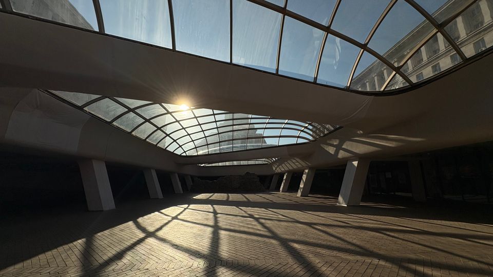 Покрита градска площ или вътрешен двор с извита стъклена конструкция на покрива и наклонени колони. Слънчевата светлина преминава през стъклата и хвърля решетъчни сенки върху павирания под.