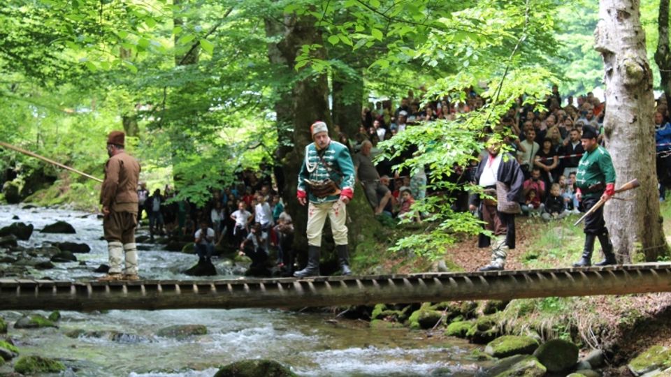 A forest setting with a wooden footbridge crossing over a shallow stream surrounded by rocks and dense green foliage. A crowd is gathered among the trees in the background, obscured by the branches.